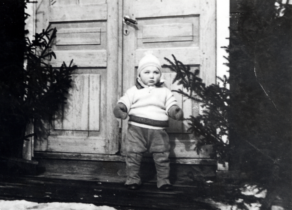 Gossen Björn Helgöstam på trappan vid Tjusta gård i Kårsta.  Björn är son till Hanna och Seth Helgöstam, gårdens ägare.  På var sida om ytterdörrarna står granar och det är snö på marken