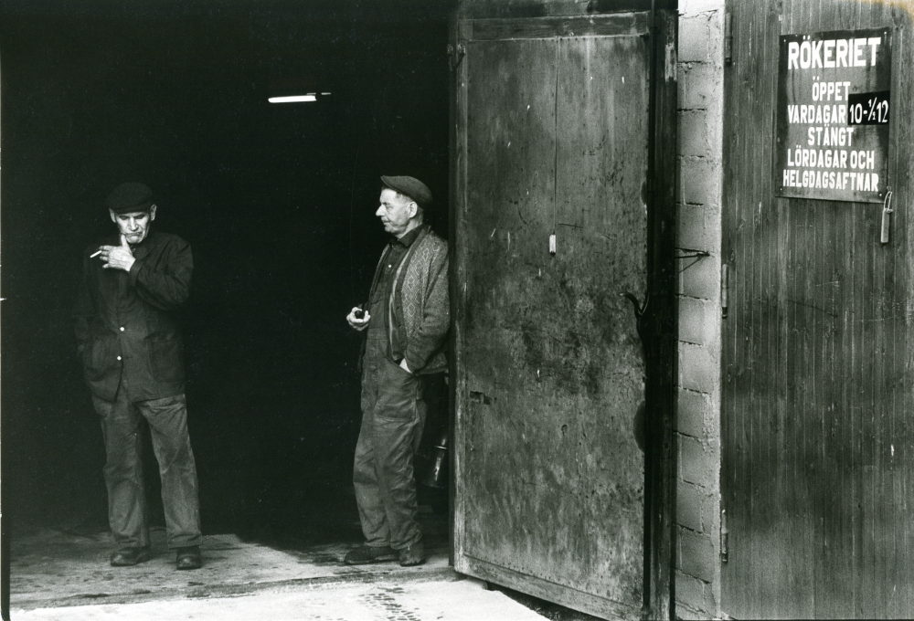 Bröderna Skyllbergs enrisrökeri i Bällsta. Bröderna Sven och Axel Skyllberg tar en paus i dörren till rökeriet. Rökeriet låg på Bällstalundsvägen. Rökeriet var i drift till slutet av 1970-talet. Senare användes lokalen till verkstad och uppställningsplats för bilar. 
•Fotografiet ingår i bildserien ”Vardagsliv i Vallentuna kommun” av Rachael Gough-Azmier & Gunilla Blomé, Haga Studios HB.
