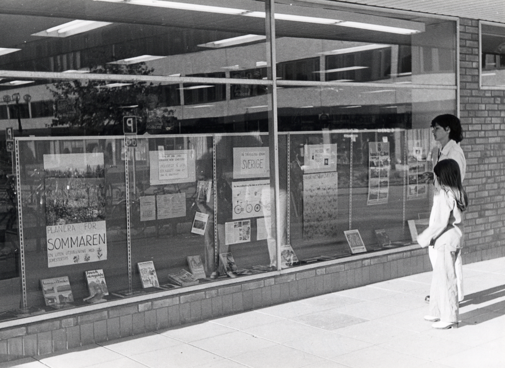 Här visar Vallentuna bibliotek en liten utställning med  semestertips av olika slag inför sommaren 1977 i Kulturfönstret i Konsumhuset, nuvarande Torghuset, Sko-Eies skyltfönster.  Kulturfönstret flyttade med till bibliotekets nya lokaler i Tärningen 1982, t v om ingången till husets kontorslokaler