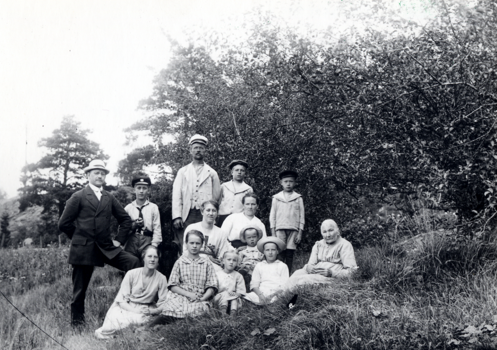 Det var förr vanligt att stockholmsfamiljer hyrde sommarnöje i vallentunatrakten. Flera år bodde fanjunkare Häger och hans familj hos trädgårdsmästare Erik Edlind på Eriksdal i Kårsta. Här har man samlats i gröngräset. Fr v fanjunkare Häger, Sven Häger, Ingeborg?, Dagmar Edlind, 1907-1979, Erik Edlind 1877-1969, Fru Häger, Oskar Edlind 1909-, fru Hilma Edlind 1882-1969, med sonen Olle Edlind 1917-1975, i knäet, framför modern sitter Svea Edlind 1912-, bredvid modern Åke Edlind 1914-, och den gamla damen är farmor Häger. Publicerad i Vallentuna kommun-nytt 13(1985): 3. Svar från Åke Edlind i Kårsta omgående
