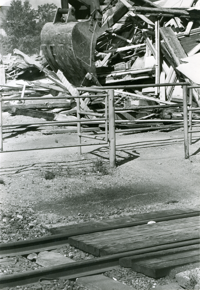 Rivning av Vallentuna stationshus pågår. Grävskopan sätter tänderna i resterna av stationshuset. I förgrunden syns övergången för gångtrafikanter.
•Fotografiet ingår i bildserien ”Vardagsliv i Vallentuna kommun” av Rachael Gough-Azmier & Gunilla Blomé, Haga Studios HB.