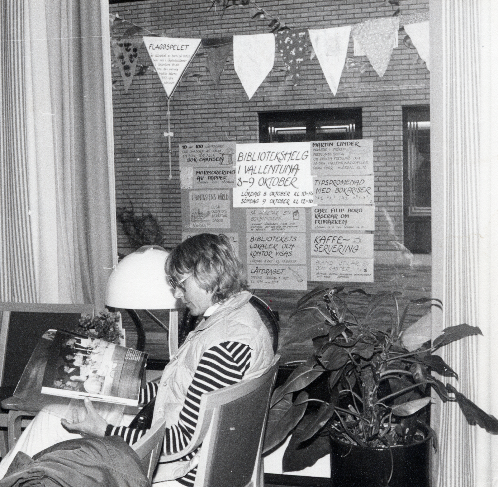 Kerstin Bark sitter i tidskriftshörnan i Vallentuna bibliotek och läser.  I fönstret hänger reklamskyltar för program och aktiviteter till boken fyller år, den tryckta bokens 500-års jubileum. Utanför fönstret skymtar flaggspel som barn tillverkat