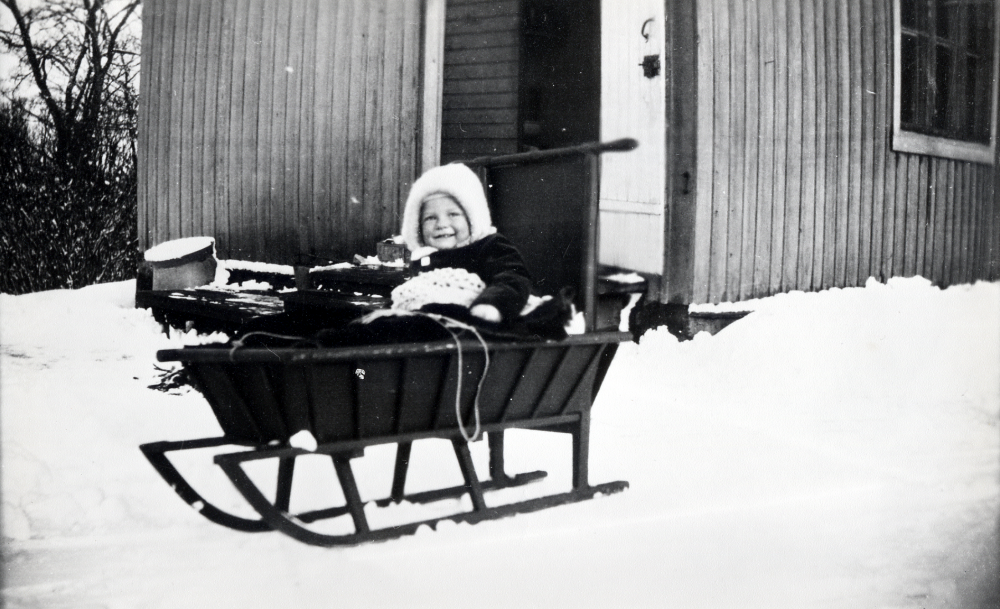 Mona Tiberg, född 1930, sitter i en kälke på gårdsplanen utanför den tillbyggda köksdelen vid Bällsta gård i Vallentuna.  Mona är dotter till Gunnel och David Tiberg. Davids syster Elsa Tiberg drev pensionatsrörelse på gården 1928-1933