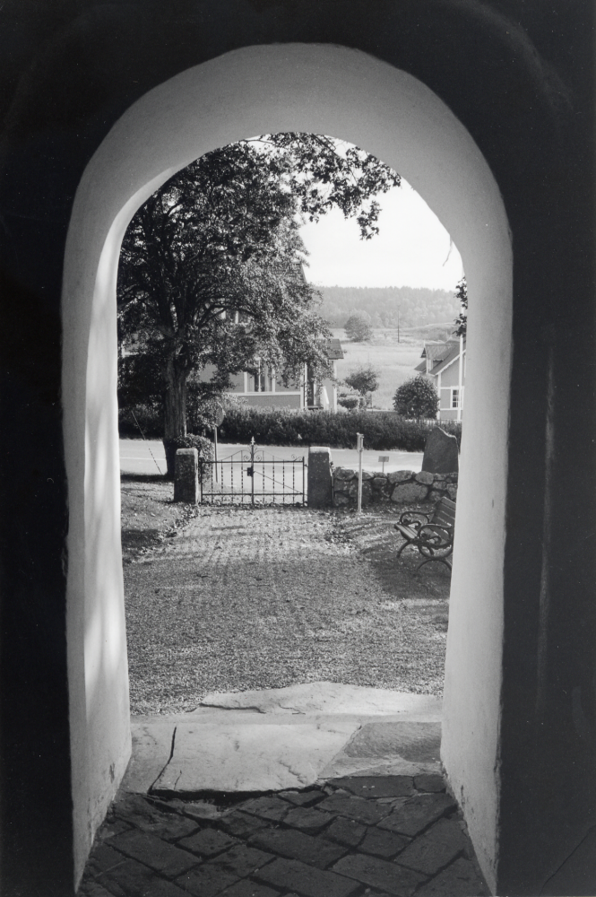 Vy söderut genom kyrkporten i Angarns kyrka.  Bostadshuset söder om kyrkan är den gamla lanthandeln Karlborg, i verksamhet till mitten av 1960-talet