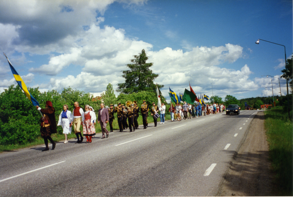 Sveriges nationaldag och svenska flaggans dag firas den 6 juni 1993. Deltagarna  marscherar på Väsbyvägen från Kommunalhusparken till Väsby kvarn med Svenska flaggan och Röda korsets flagga. Allra längst bort i bild skymtar bommarna vid järnvägsövergången.
Foto och uppgifter: Fredrik Andersson.