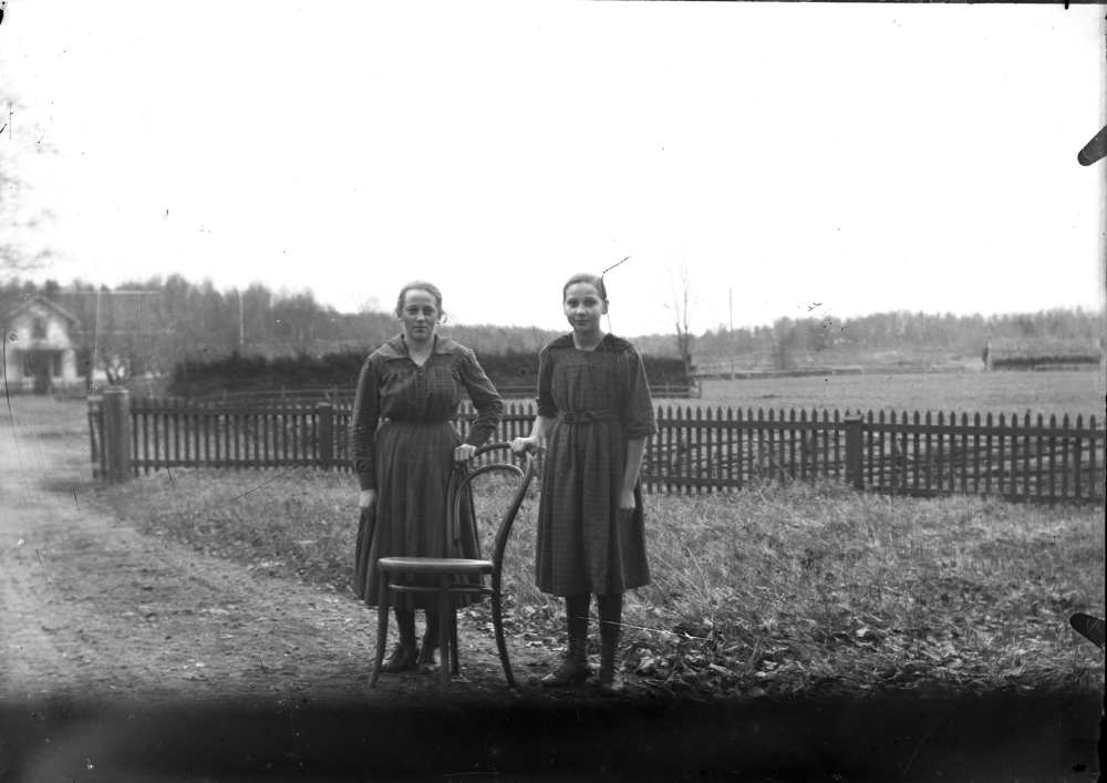 Två kvinnor och en stol, framför ett staket. Platsen är okänd. Uppgift om fotoår saknas, men troligen 1920-talet.