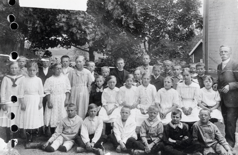 Läraren Zackeus Carlsson tillsammans med skolbarnen utanför Frösunda skola examensdagen 1919. Fotografiet skadat av hål i vänster sida. Bakre raden fr v Märta Johansson, Stig Uddenberg, Laila Lindkvist, Evert Lundkvist, Alice Helgöstam, Holger Ölund, Gunhild Åkerlind-Wredh, Gunnar Andersson, Folke Pettersson, Oscar Karlsson, okänd, Harald Wredh, Arnold Jansson, ? Uddenberg, Albin ?, Gösta Johansson, Birger Petersson, Svea Lundkvist, Wera Österberg, Inez Gustafsson-Mattson, Svea Johansson, Barbro Waldebäck, Betty Johansson, Laura Waldebäck och läraren Zackeus Waldebäck. Nedre raden fr v Sigvard Wredh, Stig Johansson, Harald Waldebäck, Holger Ljungstedt, Nils Karlsson och Erik Andersson