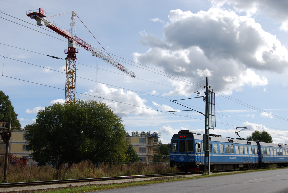 Tåget på Roslagsbanan kör norrut från Vallentuna station, sett från Banvägen. På andra sidan tågspåret syns en lyftkran vid bygget av Tegelbackens seniorbostäder.