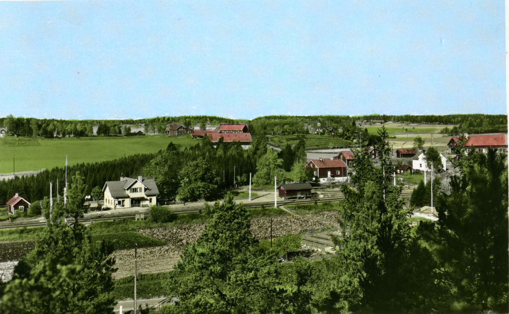 Vy över Kårsta stationsområde med Backa gårds ekonomibyggnader i bakgrunden. Det röda huset t h om stationshuset är Staffs f d affär och bakom det skymtar tornet på Backa gård mangårdsbyggnad. Samma motiv som bild 6526 men detta är handkolorerat. Vykortsfotografen Elliord Mattsson skickade ibland sina kort till Tyskland för att koloreras. Överensstämmelsen med de verkliga färgerna på hus och bilar var inte alltid total.