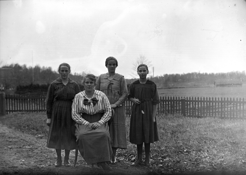 Fyra okända kvinnor står uppställda framför ett staket. Platsen är okänd.

Uppgift om fotoår saknas, men troligen första delen av 1900-talet.