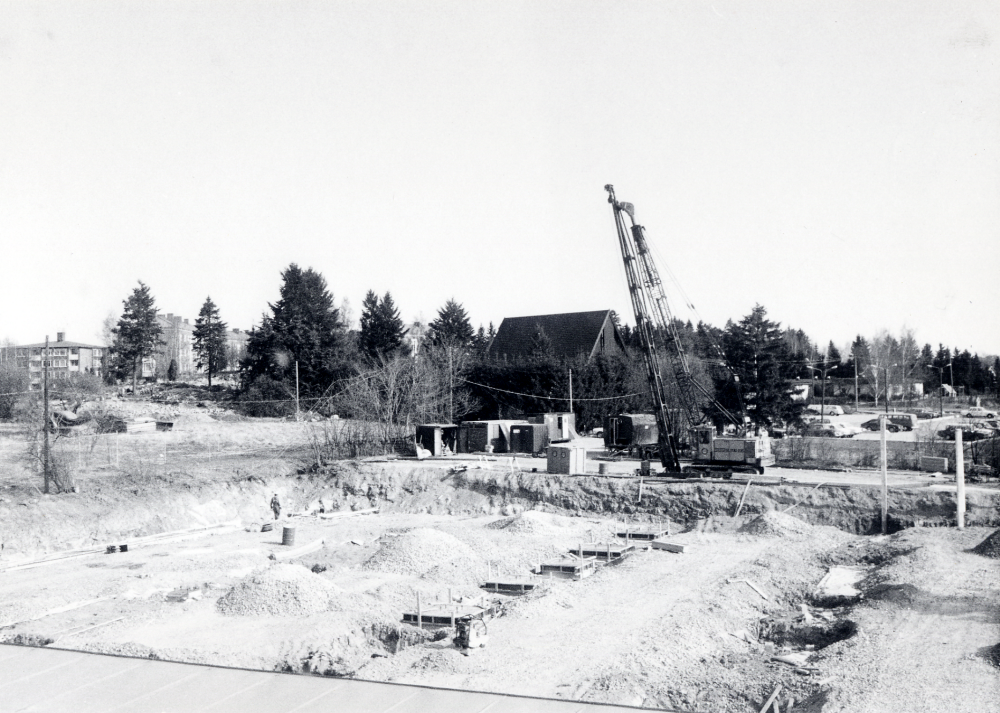 Ett nytt Domusvaruhus byggs i Vallentuna centrum norr om Konsumhuset.  Efter schaktningsarbeten för det blivande Domusgaraget  täcks marken av ett lager makadam.  Pålningsarbeten pågår för grundförstärkning  – marken är gammal sjöbotten.  Gjutningsarbeten har påbörjats.  I bakgrunden fr v syns flerfamiljshus vid Mörbyvägen, Pingstkyrkan och daghemmet Videgården