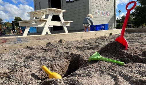 Sandlåda med spadar i sanden. Barn kan i bakgrunden ses gräva i en blå låda efter leksaker.