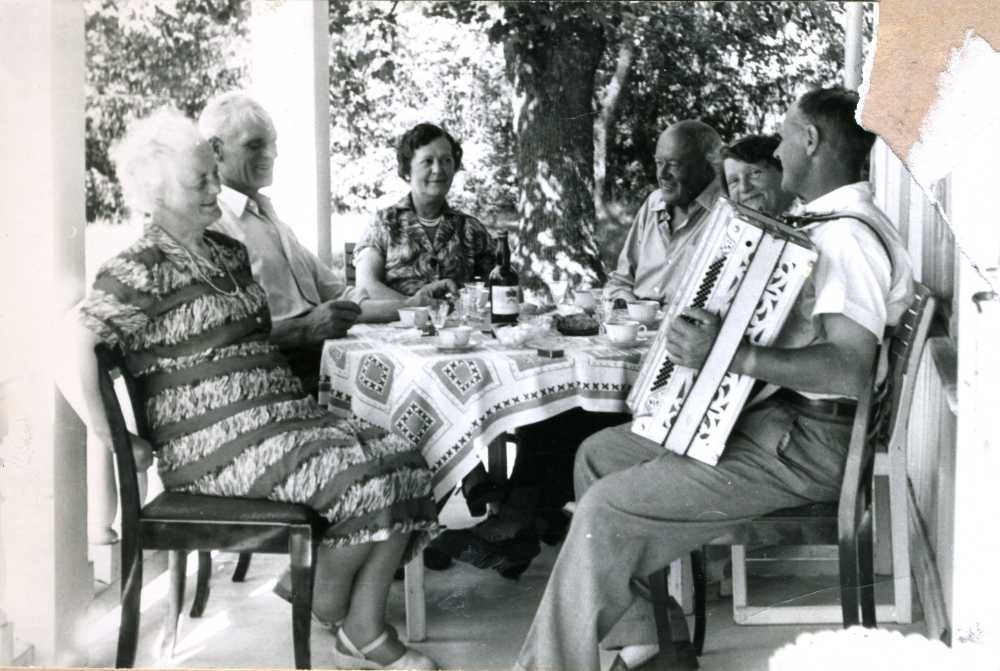 På nedre västra verandan sitter från vänster Inez och Manfred Ekholm, Adina Lind, Nils Sundmark, Vera Sundmark och Elve Berlin med dragspel. Nils och Vera Sundmark var sommargäster på Ekskogens gård. Elve hjälpte till med jordbruksarbetet på somrarna.
