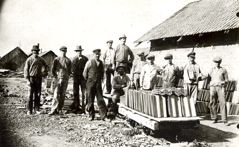 Vallentuna teglbruk uppfördes ca 1920 av fabrikör A W Lundqvist. På tegelbruket tillverkades taktegel, byggnadssten och tegelrör. Här har arbetarna samlats vid en vagn som tillhör brukets decauville-bana. Bruket var i drift fram till julen 1967 och revs 1969. Längst t v står Johansson och bredvid honom Josef Kling. Nr 3 fr v är Ejnar Wallin, Vallskog vid Björnbodavägen, där det tidigare låg ett gammalt soldattorp. Ev är det Axel Lundqvist själv som står i bildens mitt iklädd hatt