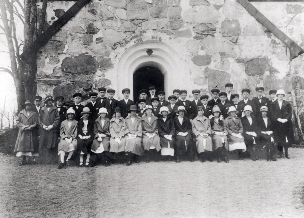 Kyrkoherde Olle Mattsson med konfirmander utanför Össeby-Garns kyrka.  Pojken vid krysset är Valter Craemer.  Ovanligt med flickor i ytterkläder och framförallt hattar i samband med fotografering vid konfirmationen