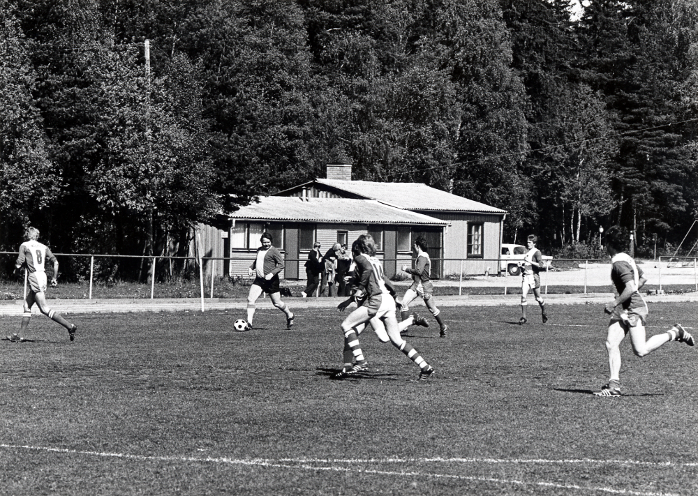 Fotbollsmatch mellan gamla VBK:are, Vallentuna bollklubb VBK fotbollslaget och 'blandat gubblag' på Vallentuna idrottsplats Den legendariske fotbolsspelaren Gösta 'Knivsta' Sandberg har bollen. Nr 8 är Lennart Holm nr 14 Kent Olsson vid huset Lennart Breimo vid bilen Mats Huppel och längst t h Lennart Jansson