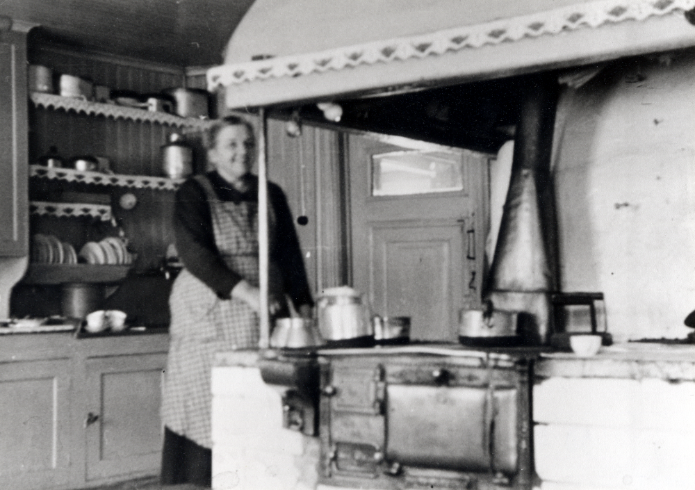 Elin Johansson, gift med gårdens ägare Emanuel Johansson, står i köket på Helgö gård vid den stora spisen.  Huvudbyggnaden brann ner till grunden 1960.  Dottern Alice har virkat  hyllremsorna