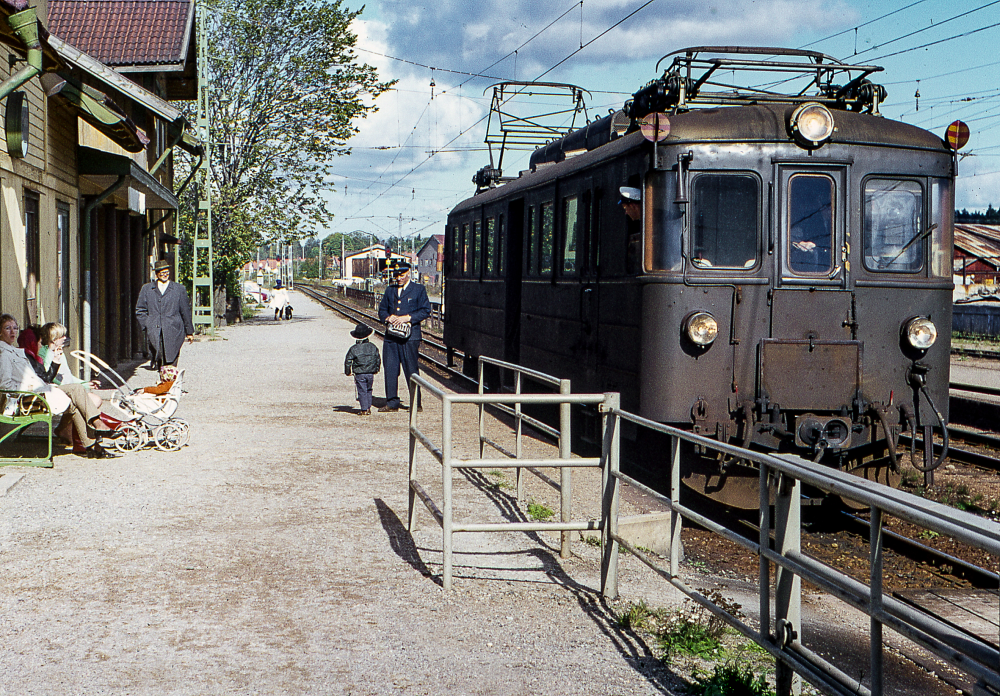 Vid Vallentuna station. Ett tåg har stannat vid stationen, konduktören står på perrongen och pratar med en litet barn. Väntande resenärer sitter på en bänk framför stationshuset.
Bildserie: Vallentuna på 1960-talet.