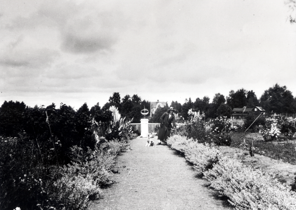 Claës Erik Schram köpte 1921 tomtmark öster om Ormsta gård för att bygga ett sommarställe.  Det fick namnet Schramsta.  Claës Erik Schram anlade en trädgård med solur, blommor och fruktträd.  Här står hustrun Marie-Louise med familjens hund vid blomrabatterna och ett solur i bakgrunden.  Bakom staketet syns Solsta, bostad för Marie-Louises bror konstnären C M Carlsson och hans familj.  T v syns Solstas hönshus