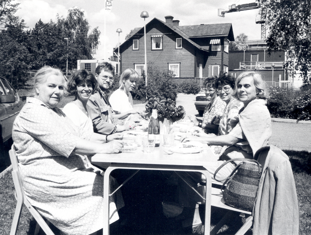 Personalen vid Vallentuna bibliotek har flyttat ut ett bord och äter lunch utomhus på gräsmattan vid Tärningens baksida. I bakgrunden syns den kvarvarande syskonvillan. Vid bordet sitter på vänster sida Inger Olsson, Lena Lundgren, Björn Svensson och Britt-Marie Thomaeus-Allen. På höger sida fr v Lena Drevemo, Eva Rahmqvist och Ingrid Ullman
