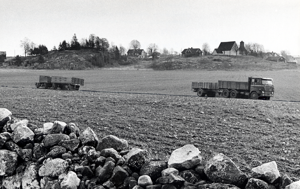 Angarns kyrka sedd från Örstafältet. Lastbilarna är monterade för att visa hur en ev vägdragning kommer att påverka landskapsbilden. Angarns kyrkby ses i bakgrunden.