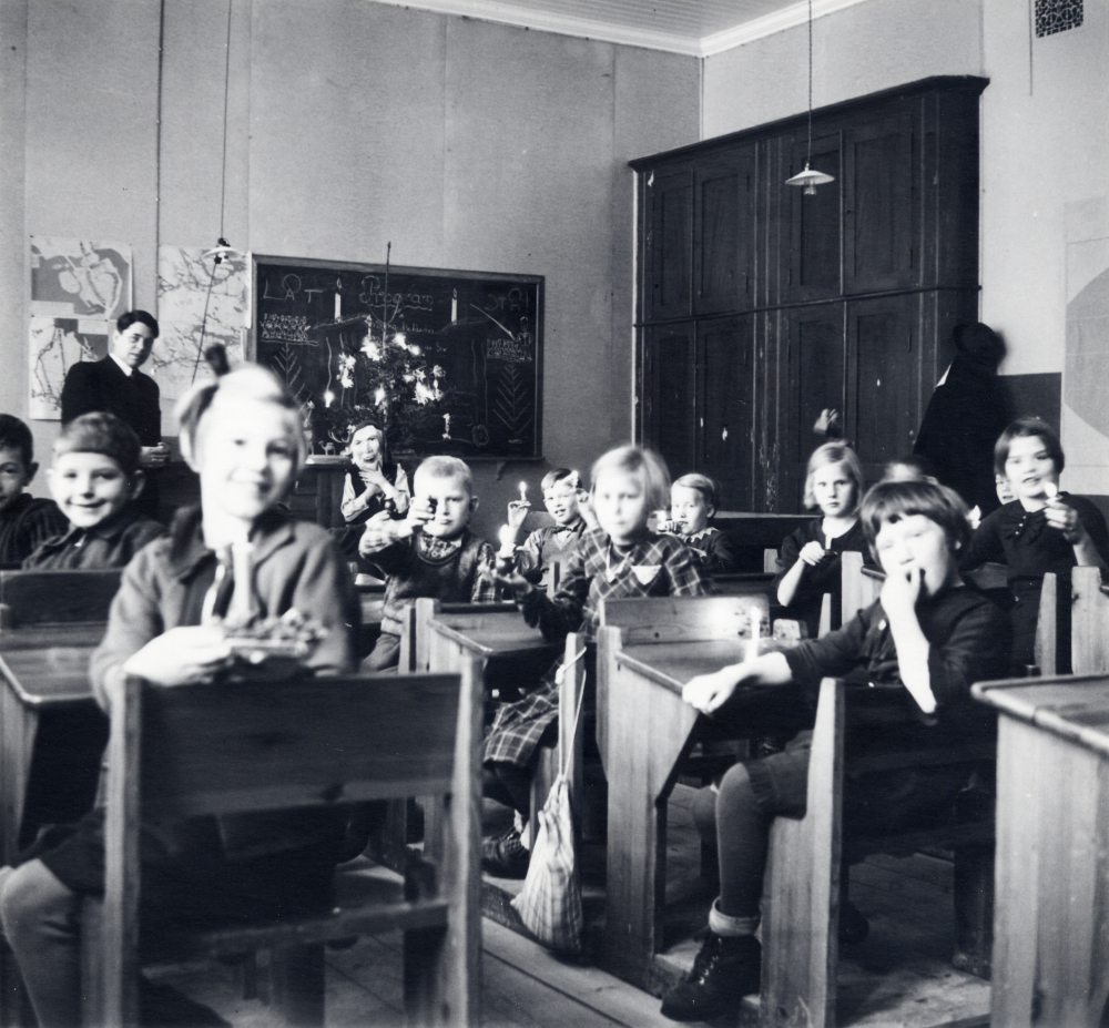 Läraren Josef Andersson tillsammans med skolbarnen i klassrummet i Vita skolan i Hjälmsta Förmodligen julavslutning Det finns julgran framme vid katedern, alla barnen har ljus på bänkarna eller i händerna, en pojke har tomtemask På svarta tavlan står program uppskrivet. Bilden har varit efterlyst i Vallentuna kommuninformation utan resultat
