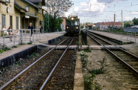 Vid Vallentuna station. Ett tåg ankommer till stationen. Väntande resenärer sitter på en bänk framför stationshuset. Huset revs 1972. Det lite mörkare huset är det gamla järnvägshotellet med restaurang som kallades Flugan. Byggnaden brann ner 1970. 
Bildserie: Vallentuna på 1960-talet.