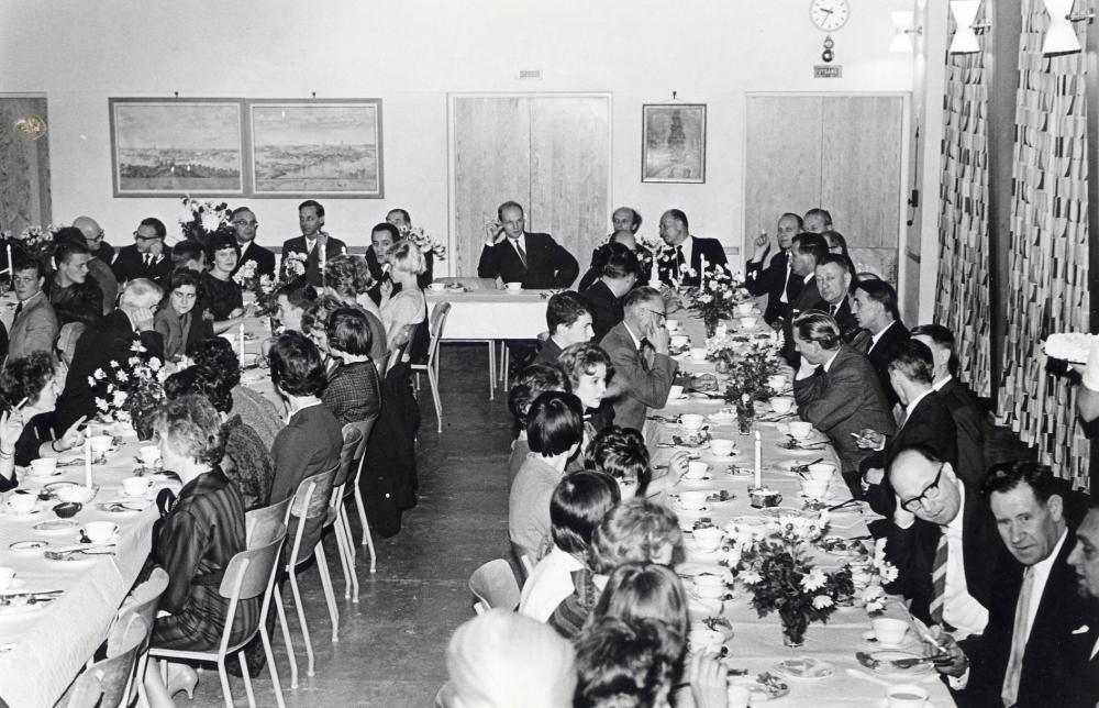 Sporthallen i Hjälmsta skola invigdes den 8 nov 1961. Här har man samlats i aulan för att äta middag efter invigningen. Vid bortet t h på höger sida sitter ingenjör Petter Nordin, elmontör K E Karlsson, socialchef Elis Pettersson, posttjänsteman Charles Pettersson, konduktör Elof Österberg, snett emot kantor Holger Hovmark. Längst bort framför dörren sitter ingenjör Diurson och arkitekt C H Bergman