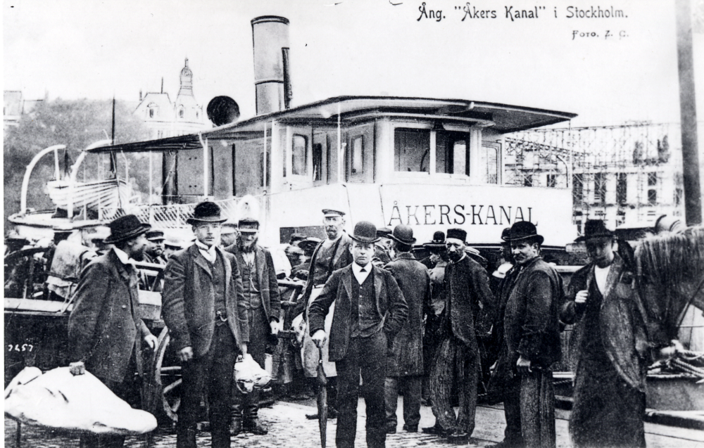 Ångfartyget Åkers kanal gick i trafik mellan Össeby-Garn och Stockholm fram till 1927 med gods och passagerare. Båten kallades Tuppen. Här ligger båten vid Nybroviken i Stockholm. På kajen finns flera män samlade.