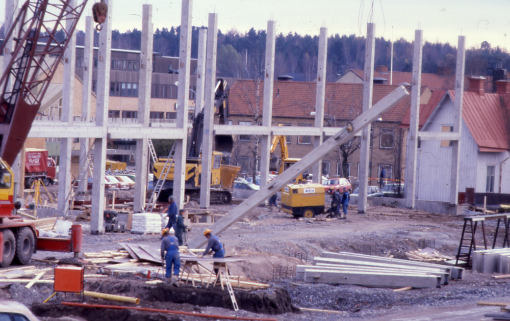 Stommen till det som kommer att bli Förvaltningshuset Tärningen har börjat resa sig över marken. I bakgrunden syns, från vänster, Torghuset, Tuna huset, gamla kommunalhuset och villan där affären Kära Barn hade sin verksamhet.