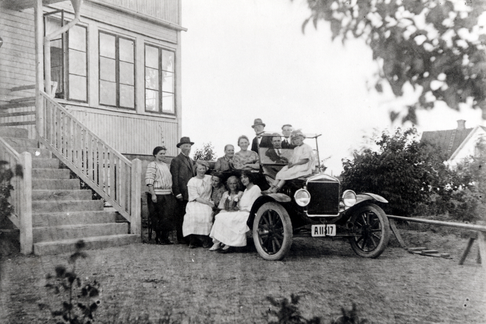 Personbil utanför Villa Edsberg på Allévägen.  Personerna vid bilen fr v är Ingeborg Gustafsson, gift Svedberg, skomakare K P Gustafsson som bodde i Villa Edsberg, i bilen okända personer utom Ernst Gustafsson längst t h. På fotsteget sitter fr v Rut Gustafsson gift med Cyrus, Mimmi Gustafsson gift Palmblad, May Svedberg och Beda Gustafsson. På motorhuven sitter Alli Svedberg.
