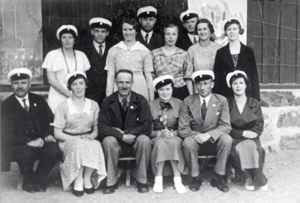 Vallentuna hembygdskör  samlad utanför  Hjälmstaskolan  Bakre raden fr v  Edit Lindström   ? Tanders   Tora Krieg   Josef Andersson  okänd,  Arne Lindström   Aina Bornholm  och  Aina Andersson  Främre raden fr v okänd,  Gerda Groth   Erik Jonason   Gun ?   Rickard Jansson  och  Maj Linder
