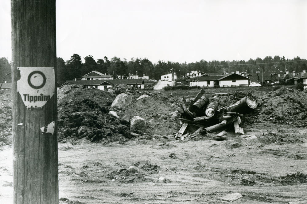Vägen mot Molnby med Snapptuna i bakgrunden. Det står "Tippning förbjuden" på en trasig skylt uppsatt på stolpen till vänster. Gamla oljefat och plank är dumpade på marken.
•Fotografiet ingår i bildserien ”Vardagsliv i Vallentuna kommun” av Rachael Gough-Azmier & Gunilla Blomé, Haga Studios HB.