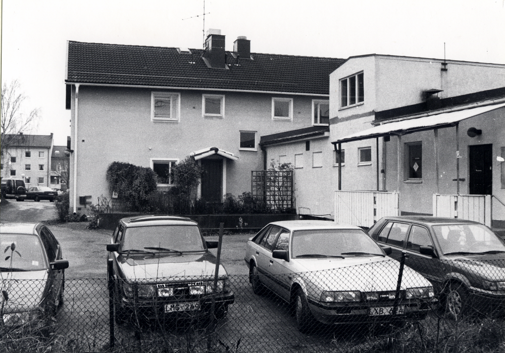 Alla fastigheterna på Allévägens norra sida mellan centrum och Mörbyvägen skall rivas.  Här syns baksidan av Handelsbankens hus. Succé Färg och Tapetlager har haft sina lokaler i det vinkelställda huset men har redan flyttat till Okvista industriområde.  Tre bilar står parkerade på gården