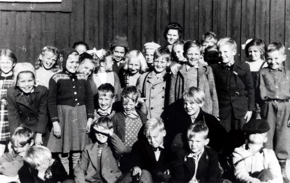 Lärarinnan Kerstin Lundqvist med skolbarnen utanför Röda skolan.  I övre raden stående fr v Britt?, Britt Öberg, Siv Pettersson (Jansson,) Gun Hallenberg, Siv?, Ingrid Eriksson (Hammarstedt), Kristina Strid, Britt-Marie?, Maj Lutteman, Lars Bornholm, Maj-Britt Öberg, Lennart Söderström, Göran Ekström (Iveberg), Birgit Kallberg, Hans Andersson, Evy Lööf och Björn Skogström.  Rad två på knä fr v Karin Dahl, Hans Johansson (Lifsten), Börje Hofgren och Yngve Tillborg.  Sittande längst ner fr v Björn Uhrenius, Harry?, Ulf Sundborg, Olle Bergström, Staffan Lindhé och Christer Viwanger