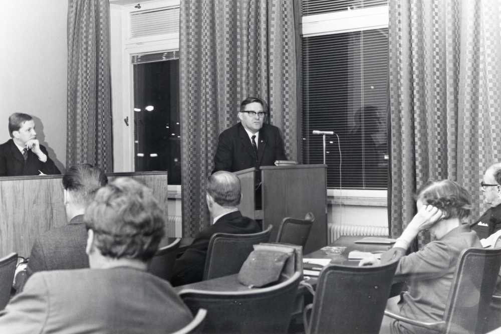 Kommunfullmäktige sammanträder i sessionssalen i kommunalhuset. På podiet sitter t v Ulf Berglund. I talarstolen står Bertil Tillborg. Damen med handen vid örat är Karin Edman.