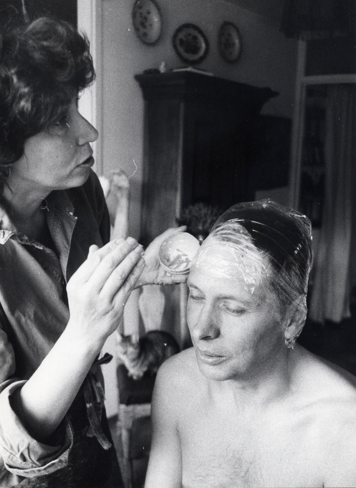 Eva Rahmqvist gjuter av ansiktet på Gunnar Hillbo med hjälp av gipsbindor. Masken skall användas till ansiktet på en snickare på Läckö slotts sommarutställning Trä 1987. Ansiktet smörjes in med vaselin och gipsbindor, klippta i små bitar och doppade i vatten, läggs på ansiktet. Efter en stund dras gipset bort och Eva modellerar sedan ett huvud med utgångspunkt från det gjutna ansiktet. Här stryker Eva in vaselin på Gunnars ansikte, sedan håret täckts med plast. Foto från Eva Rahmqvists hem, Bällstavägen 59 C.