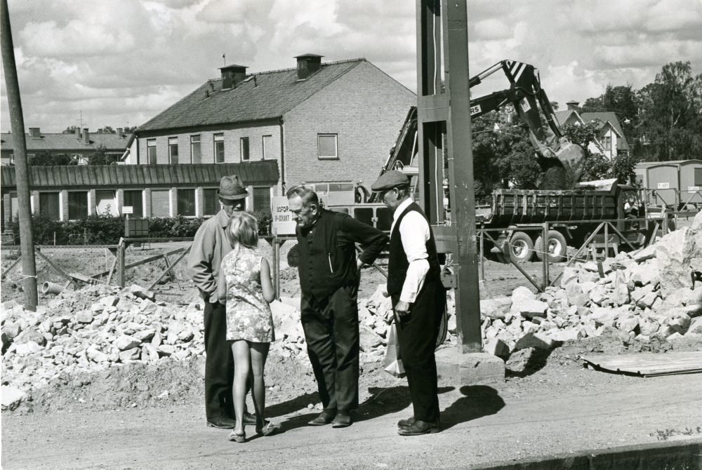 Rivning av Vallentuna stationshus pågår. Helge Almqvist, före detta lokförare E Österberg och Albin Källström pratar med en ung flicka på perrongen framför rivningsmassorna.
•Fotografiet ingår i bildserien ”Vardagsliv i Vallentuna kommun” av Rachael Gough-Azmier & Gunilla Blomé, Haga Studios HB.
