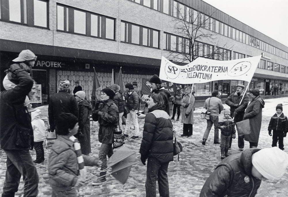 Sveriges arbetareparti m fl socialistiska partier i Vallentuna ordnar första maj-demonstration med samling bakom Centrumbio för att tåga till Tuna torg. Här har man kommit fram till Tuna torg. Möjligen har man samlats först vid Sörgårdstorget för att tåga Allévägen ner mot centrum. En banderoll med texten SAP Socialdemokraterna i VAllentuna hålles i mitten av Karl Erik Nilson