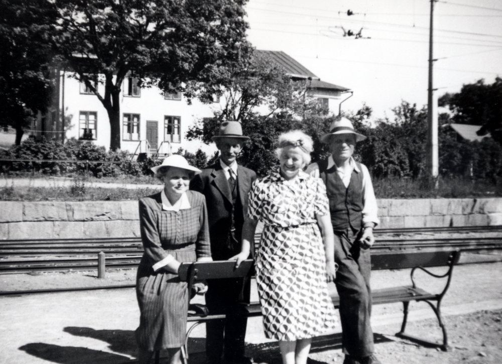 Ekskogens järnvägsstation med Charlottenberg i bakgrunden. På perrongen står fr v sömnadslärarinnan Svea Nygren, lantbrukaren Klas Nygren, Inez och Manfred Ekholm, ägare till Ekskogens gård under åren 1947-1965. Fotograf är deras adoptivson Timo, som kom till familjen som finskt krigsbarn under andra världskriget. I Charlottenberg fanns en handelsbod i bottenvåningen och en samlingslokal i övervåningen. I huset låg också Ekskogens postkontor