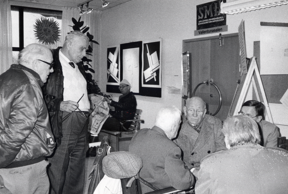 Sme-Kalle   K S Karlsson  besöker  Vallentuna bibliotek  och tittar på  utställningen   Smia  i Vallentunasalen, arrangerad av  Hans Lustig  och  Upplandsmuseet  I bildens mitt i rullstol sitter Sme-Kalle själv och samtalar fr v med fr v  Erik Sundborg,   "Plåt-Janne" Jansson,   Gustav Nilsson   Halvar Näslund  med ryggen mot kameran och initiativtagare till besöket, samt  Erold Sandquist