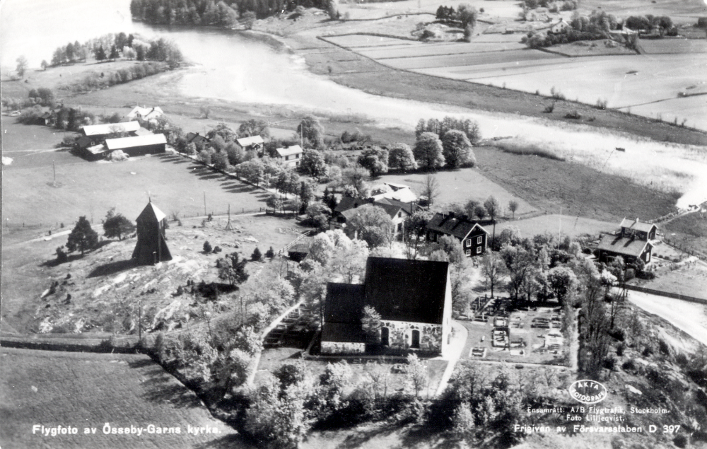 Flygfoto  över  Össeby-Garns kyrkby, Lilla Garn,  med kyrkan i förgrunden och  Garnsviken  längst upp t v med  Husaån.  Ångfartyget Åkers Kanal trafikerade sträckan Brottby- Stockholm. Fartyget vände mitt för nuvarande brandstationen och bensinstationen, där det fortfarande finns rester av bryggan. Ån lär ha muddrats bredare här för att båten skulle få plats att vända. Uppgift om fotoår saknas, men troligen första delen av 1900-talet.