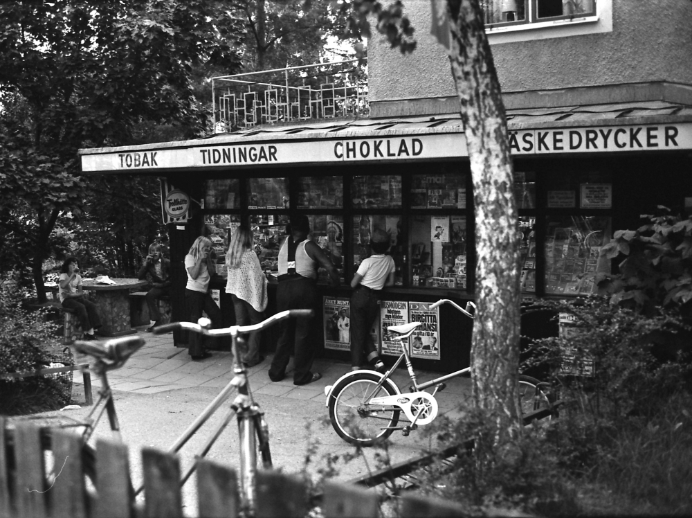 Kunder utanför Bällsta kiosk vid Bällsta station. Tobak, tidningar, choklad och läskedrycker står tryckt i versaler på kiosktakets front.

•Fotografiet ingår i bildserien ”Vardagsliv i Vallentuna kommun” av Rachael Gough-Azmier & Gunilla Blomé, Haga Studios HB.
