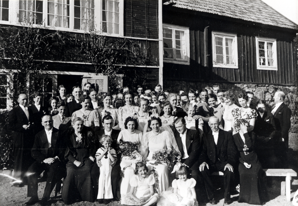 Dubbelbröllop på Helgö gård.  Elin och Emanuel Johansson på Helgö gifter bort två av sina döttrar. Utanför gården är bröllopsgästerna samlade.  Främre raden fr v Oscar Andersson, Elin Johansson, brudgummen Henning Linde, bruden Signe Helgöstam, bruden Margit Helgöstam, brudgummen Holger Andersson, Emanuel Johansson och Lisen Andersson.  Flickorna är Stina och Karin Céwe.  Pojken är Lars Helgöstam