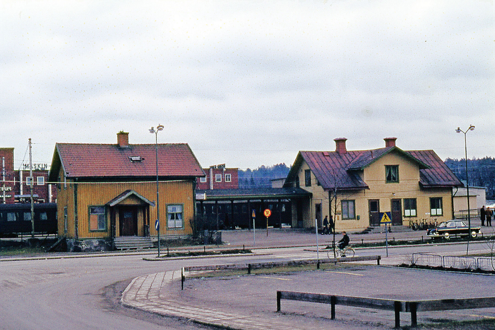 Det lite mörkare huset till vänster i bilden är det före detta järnvägshotellet med restaurang som kallades Flugan. byggnaden brann ner 1970. Det ljusare huset till höger är Vallentuna järnvägsstation som revs 1972. I bakgrunden syns Tellus maskins lokaler. Färgbild.