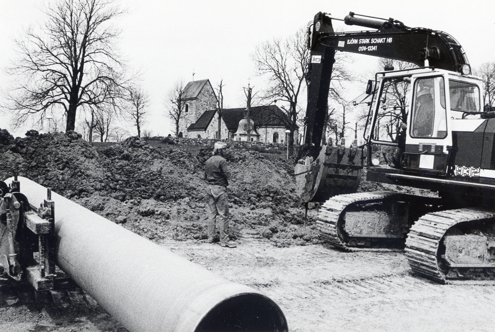 Anläggningsarbeten av Vallentuna värmeverk AB nedanför Vallentuna kyrka för rör till den nya sjövärmen från Vallentunasjön. En grävskopa gräver på åkermarken.  Stora rör skall grävas ner för att forsla det uppvärmda vatten till värmeverket. Vallentuna kyrka syns i bildens mitt