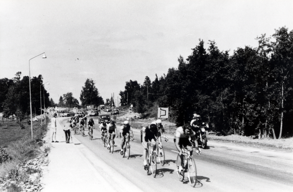 Cykeltävlingen 'sex-dagars' passerar Vallentuna 1973. Här kommer en klunga cyklister på Stockholmsvägen på väg söderut. I bakgrunden syns en omledning av trafiken på Stockholmsvägen för bygget av en gångtunnel under vägen