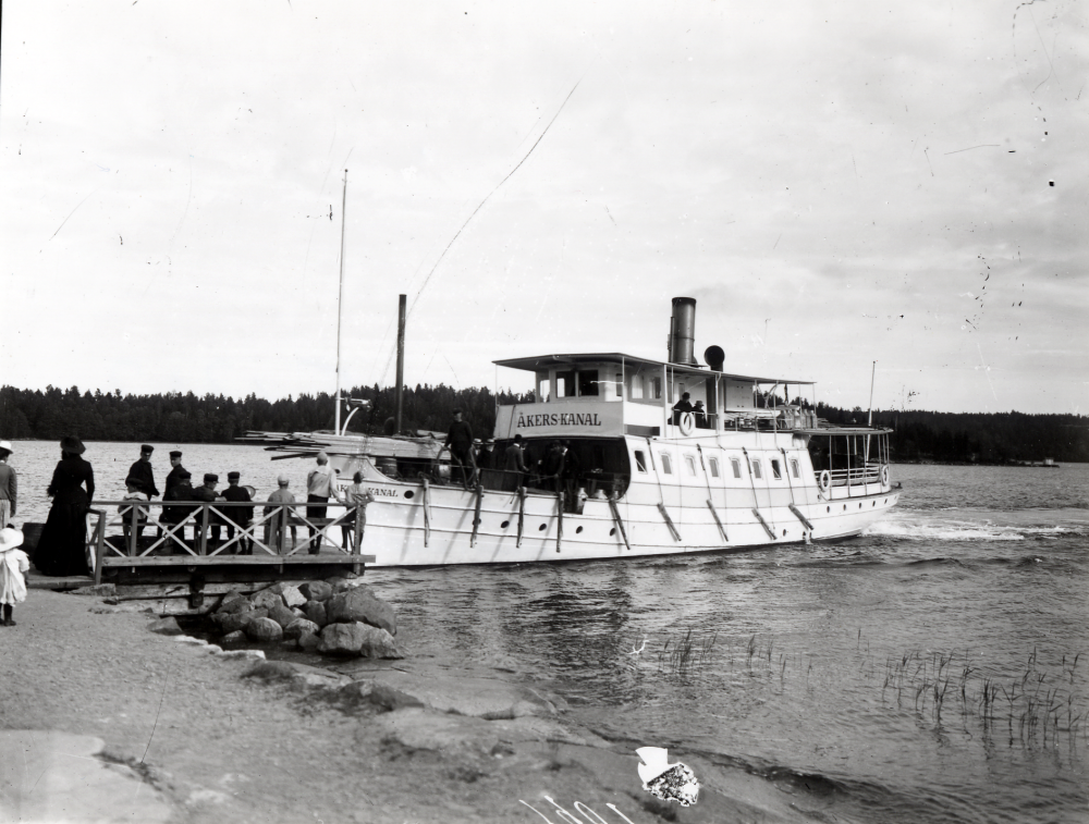 Ångfartyget Åkers Kanal vid en brygga mellan Brottby i Össeby-Garn och Stockholm. På främre däck ligger brädor. Uppgift om fotoår saknas, men troligen första delen av 1900-talet.