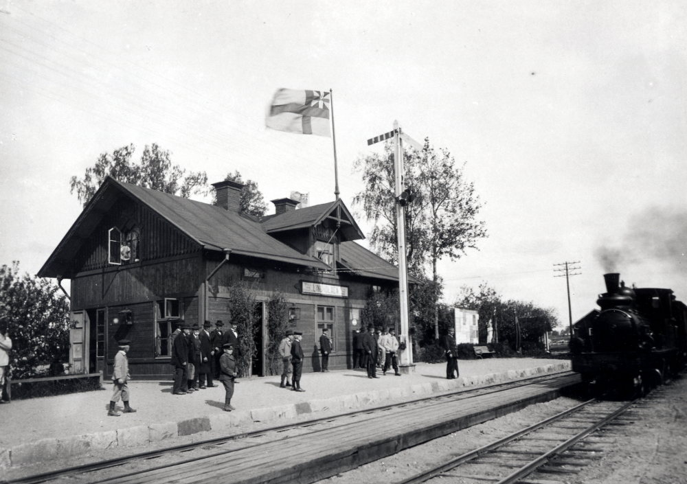I det öppna fönstret står Margit Oppermann , senare lärarinna i Vallentuna. Hennes far Henrik Oppermann var stationsinspektor på järnvägsstationen. Eventuellt firas 12 maj, Gustav Vasas födelsedag, med lövat stationshus? Årtal tveksamt, Margit föddes några år senare - ett syskon i fönstret? Årtal 1902 står skrivet på samma motiv av sämre kvalitet. Roslagsbanans tåglinje Stockholm - Rimbo invigdes 1885.