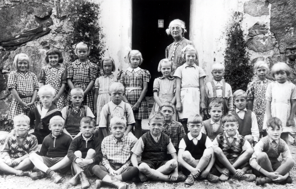 Skolfotografi med lärarinnan Nanny Skoglund och skolbarnen i Össeby-Garns småskola utanför ingången till kyrkan.  I övre raden fr v Inga Johansson, Maj-Lis Eklund, Vivian Kraemer, Kerstin Balk, Inez Jansson, Ulla Franzén, Vivian Alstermark, Rune Jansson, Ella Gustavsson och okänd.  På knä fr v Karl-Erik Johansson, Ingemar Wallbom, Hasse Gustavsson, okänd, Fritz Johansson och Tord Lundberg.  Sittande fr v Rolf Degling, Bengt Karlsson, okänd, Ture Rödin, Arne Stjärnholm, Per Lundin?, Alf Eklund och okänd.